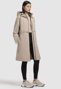 Beige waterdichte jas met een capuchon, een rits aan de voorkant en zijzakken. Draag het over een lichte top en zwarte legging, gecombineerd met beige sneakers.