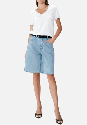 Femme portant un haut blanc à manches courtes en V, un short en jean bleu clair longueur genou avec une ceinture noire et des talons ouverts noirs.