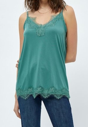 Frau trägt ein türkisfarbenes Camisole mit Spitzenbesatz an Ausschnitt und Saum, kombiniert mit dunkelblauen Jeans, vor schlichtem hellem Hintergrund.