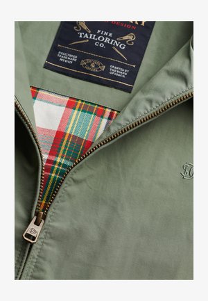 Groene jas met rits van stof, voorzien van een geruite voering in rood, geel, blauw en wit. Label met logo en merkinformatie. Ritsen van bronskleurig metaal.