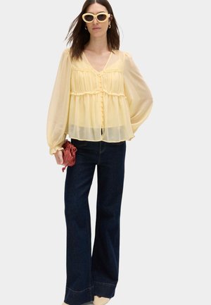 Vrouw draagt een gele doorschijnende blouse met knopen, donkere wijd uitlopende jeans, beige schoenen, crèmekleurige zonnebril en houdt een kleine bruine tas vast.