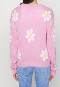 Bakifrån av en person som bär en rosa tröja med vita och gula tusenskönsmönster och ljusblå jeans.