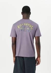 Lilla bomullst-skjorte med grafisk design på baksiden med teksten "BILLABONG", blomsterakcenter og bølger. Korte ermer, rund hals.