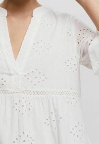 Vit linnklänning med V-ringning, med hålmönster och broderie anglaise-accenter. Designad med en lös passform och uppvikta ärmar.