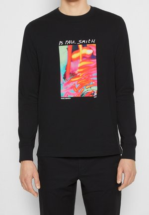 Camisa negra de manga larga con un gráfico abstracto colorido y el texto "PS Paul Smith" y "Rave Waves 1991" en el pecho, usada por una persona.