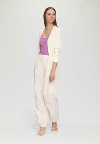 Leichter Creme-Cardigan über einem rosa gerippten Tanktop, kombiniert mit lockeren beigen Cargo-Hosen. Einfache Sandalen runden das Outfit ab.