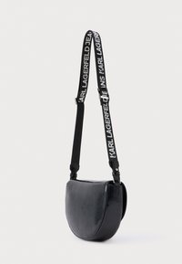 Černá kabelka přes rameno s zakřiveným tvarem, vyrobená z lesklého materiálu. Má široký popruh s bílým nápisem "KARL LAGERFELD".