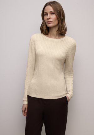 Cremefarbener, gerippter Strickpullover mit langen Ärmeln und rundem Ausschnitt, kombiniert mit dunkelbraunen Hosen. Weiche Textur und figurbetontes Design.