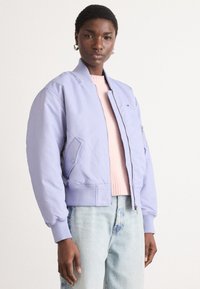 Svijetlo lavanda bomber jakna s rebrastim manžetama i pojasom. Ima prednji zip i bočne džepove. Nosena preko svijetlo ružičastog duksera i plavih traperica.