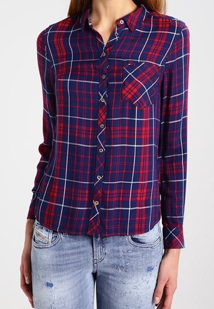 Vrouw draagt een rood- en marineblauw geruit overhemd met lange mouwen en een borstzak, gecombineerd met lichtgewassen distressed spijkerbroek.