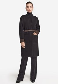 Schwarzer langer Wollmantel mit Schalkragen, braunen Ärmelabschlüssen, Seitentaschen und einem braunen Gürtel. Das Model trägt schwarze Hosen und Schuhe.