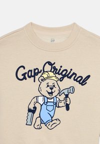 Αρκούδα που χαμογελά φοράει κίτρινο κράνος ασφαλείας και μπλε ολόσωμη φόρμα, κρατώντας πριόνι και σφυρί, με το κείμενο «Gap Original» από πάνω σε μπεζ ύφασμα.