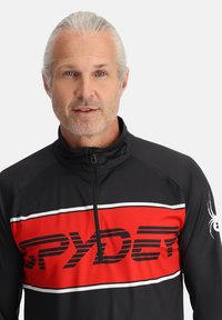 Giacca nera con zip caratterizzata da un grande pannello rosso con strisce bianche, che presenta la scritta "SPYDER" in stampa nera, con un logo a forma di ragno sulla manica.