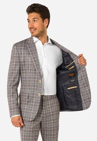 Traje a cuadros marrón claro con cierre de dos botones, botones beige y dos bolsillos interiores. Llevado sobre una camisa blanca.