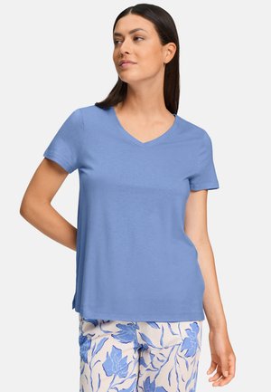 Donna con maglietta azzurro chiaro a maniche corte e scollo a V e pantaloni bianchi con motivi floreali blu, in piedi con una mano dietro la schiena.