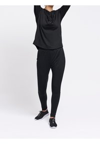 Osoba w czarnej bluzie z kapturem, czarnych dopasowanych dresach i czarnych trampkach z białymi podeszwami, stojąca na tle białego tła.