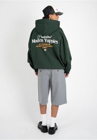 Personne portant un sweat à capuche oversize vert foncé avec l'inscription « Prohibited Modern Yuppies », un short gris à fines rayures, des chaussettes blanches et des chaussures noires, vue de dos.