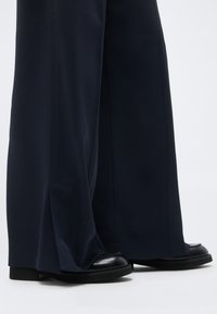 Μαύρο παντελόνι με φαρδιά μπατζάκια που καλύπτει εν μέρει μαύρα δερμάτινα παπούτσια, απεικονίζεται σε ανοιχτό φόντο.