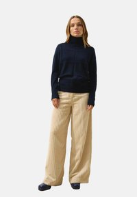 Maglione a collo alto blu navy con texture a coste, abbinato a pantaloni in corduroy beige a gamba larga. Il modello indossa scarpe scure e posa in piedi.
