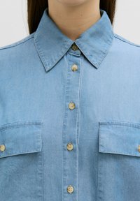 Helles, blaues Denim-Hemd mit spitzem Kragen, Knopfleiste, zwei Brusttaschen und Kunststoffknöpfen mit glatter Textur.