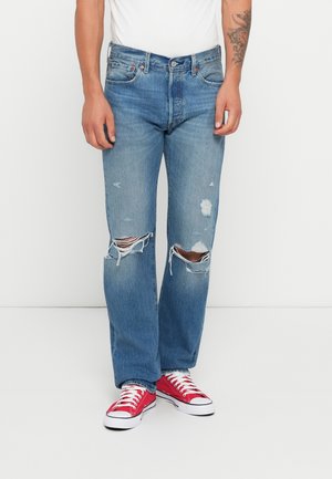 Jeans in denim chiaro sbiadito con grandi strappi sulle ginocchia, taglio dritto e un leggero effetto sfumato; abbinati a sneakers in tela rosse.