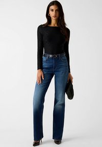 Schwarzes Langarmshirt mit strukturiertem Muster, kombiniert mit hoch taillierten blauen Jeans und schwarzen Absätzen, hält eine schwarze Clutch.
