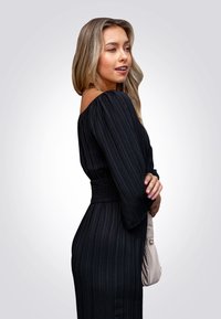Vestido negro de canalé con escote off-shoulder y mangas largas, que presenta una cintura fruncida. Se sostiene un bolso de color claro contra el costado.