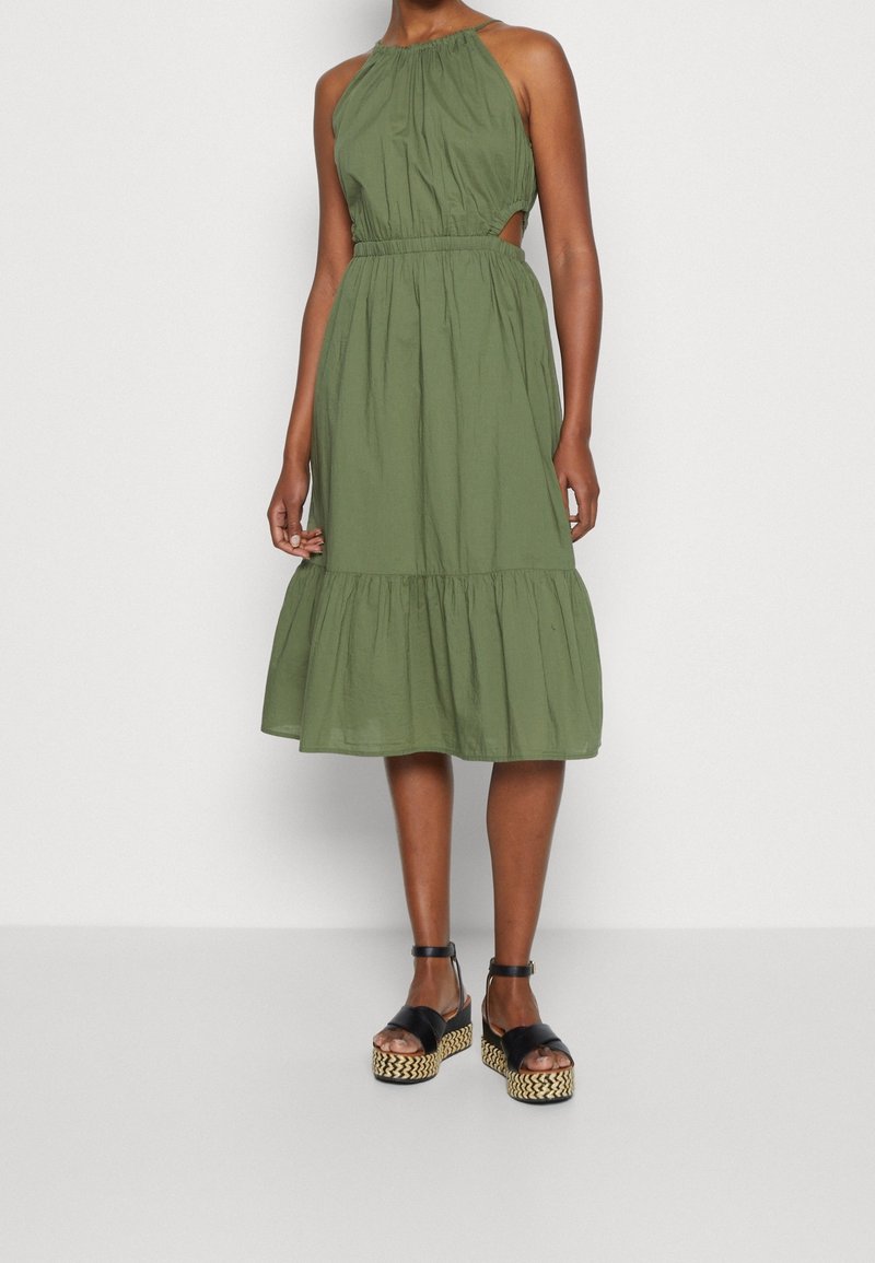 Femme portant une robe midi sans manches vert olive avec une taille froncée et des sandales compensées noires sur un fond clair uni.