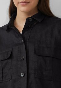 Zwarte linnen blouse met een kraag, voorzien van twee voorzakken en donkere knopen. De stof heeft een gestructureerde uitstraling en een ontspannen pasvorm.