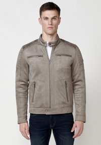 Jachetă din suede gri deschis, cu guler înalt, accente ribbate pe umeri, buzunare cu fermoar și un design ajustat. Purtată peste un tricou alb.
