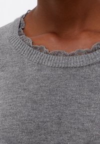 Primo piano del collo di un maglione grigio a maglia con bordatura in pizzo nero smerlato su una persona dalla carnagione scura.