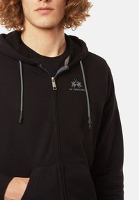 Sudadera con cremallera negra que cuenta con un bolsillo frontal, cordones grises y un logo con dos figuras, confeccionada en una tela suave.