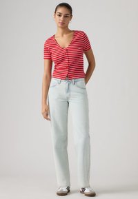 Κοντομάνικη μπλούζα με κόκκινες και λευκές ρίγες και κουμπιά, συνδυασμένη με ανοιχτό μπλε τζιν παντελόνι ίσιας γραμμής και μαύρα και λευκά αθλητικά παπούτσια.