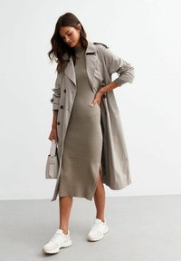 Un trench coat beige indossato sopra un vestito verde oliva a coste con uno spacco laterale. Sneakers bianche e una piccola borsa bianca completano il look.
