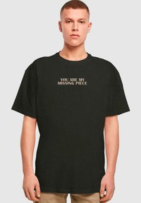 Sort bomulds T-shirt med korte ærmer, der har teksten "DU ER MIT MANGLENDE STYKKE" i en lys skrifttype centreret på brystet.