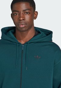 Σκούρο τιρκουάζ φούτερ με φερμουάρ και ρυθμιζόμενο κορδόνι, μαύρο λογότυπο στο στήθος, μαλακό ύφασμα και ριμπ ρίγες στους καρπούς και το στρίφωμα.