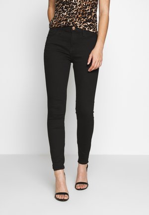 Femme portant un jean skinny noir, un top sans manches imprimé léopard et des sandales noires à talons ouverts, debout sur un fond blanc.