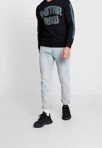 Černá mikina s velkými nápisy "G-STAR RAW", modré akcenty na rukávech. Světle modré džíny, s ohrnutými nohavicemi, v kombinaci s černými teniskami.