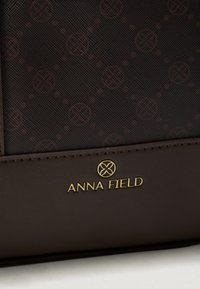 Hnědá syntetická kabelka s texturovaným povrchem a opakujícím se vzorem loga ve tmavě červené barvě. Zlatě ražené jméno značky ve spodní části.