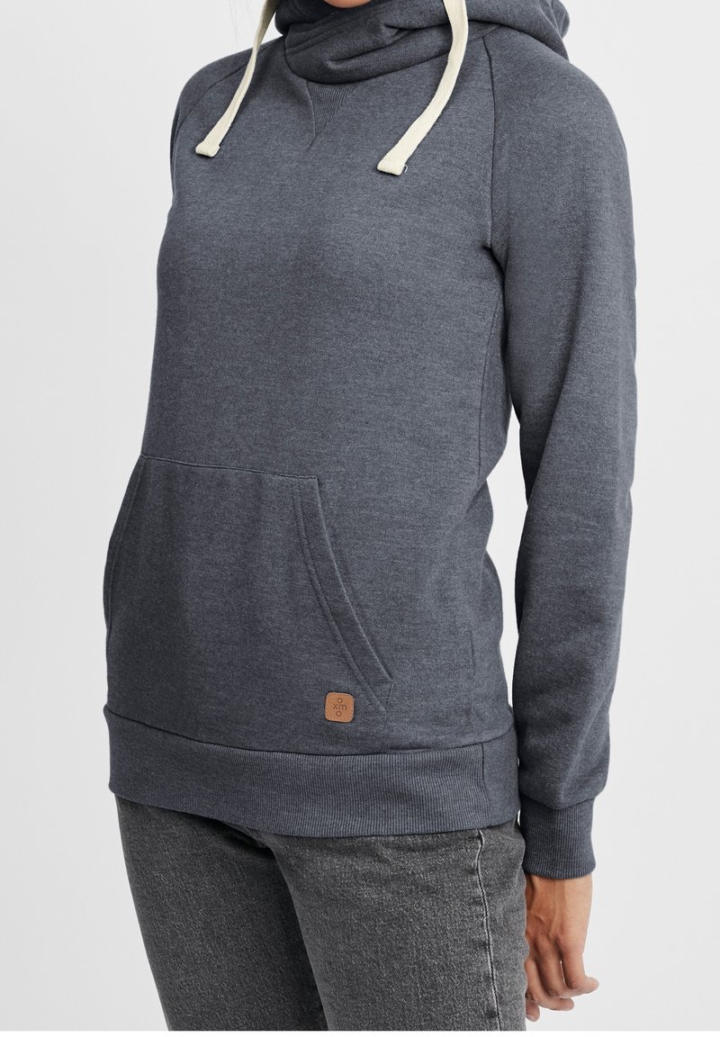 Personne portant un sweat à capuche gris foncé avec une poche frontale et des cordons de serrage crème, assorti à un jean gris délavé, sur un fond uni.