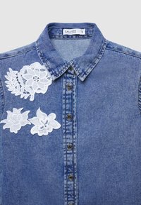 Camicia in denim con lavaggio blu, colletto classico, chiusura frontale con bottoni e motivi floreali ricamati in bianco sulla spalla sinistra e sul petto.