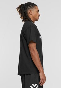 Sort bomulds t-shirt med korte ærmer, afslappet pasform, med hvid tekstdesign over fronten. Båret med sorte mesh shorts.
