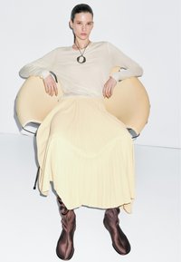 Vrouw draagt een crèmekleurige top met lange mouwen, bijpassende geplooide rok, bruine hoge laarzen en een ronde hangersketting, zittend op een moderne beige stoel.