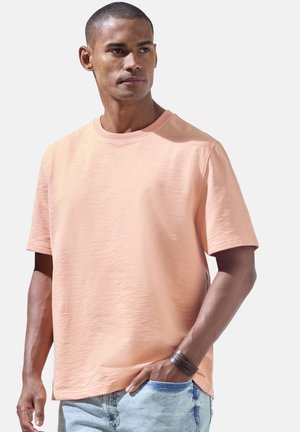 Mann in einem hellen pfirsichfarbenen T-Shirt mit kurzen Ärmeln und hellblauen Jeans, steht mit der rechten Hand in der Tasche vor einem einfarbigen Hintergrund.