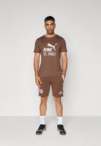 Conjunto deportivo marrón con una camiseta que presenta un gráfico blanco "KING ST. PAULI" y pantalones cortos a juego con detalles laterales y logotipos.