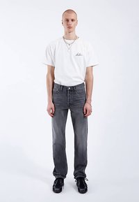 T-shirt bianca a maniche corte con logo nero, abbinata a jeans grigio scuro a gamba dritta e scarpe nere. Design semplice, senza motivi o accenti.