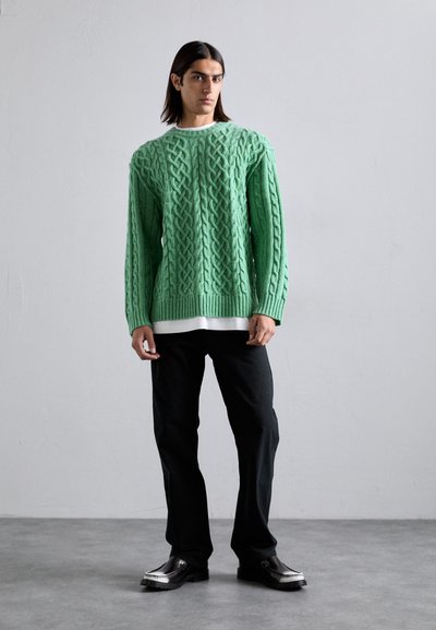 Suéter verde de punto con un grueso dobladillo acanalado, combinado con una camisa blanca. Pantalones negros y zapatos negros brillantes completan el conjunto.
