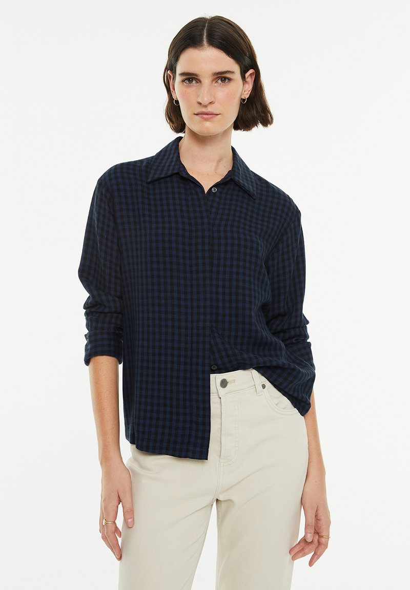 Camisa de cuadros azul marino con botones, de manga larga y corte relajado. Cuenta con un collar y puños de manga enrollados, combinada con pantalones beige.
