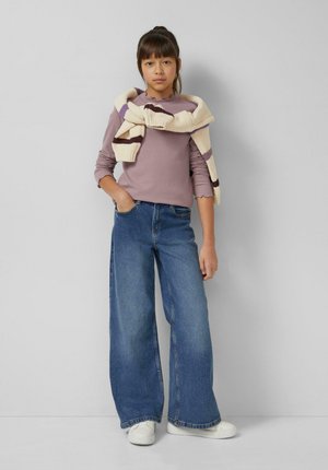 Lichtpaars geribbeld shirt met lange mouwen en gegolfde boorden, gedragen met blauwe wijde jeans en een crèmekleurige gestreepte gebreide trui over de schouders gedrapeerd.