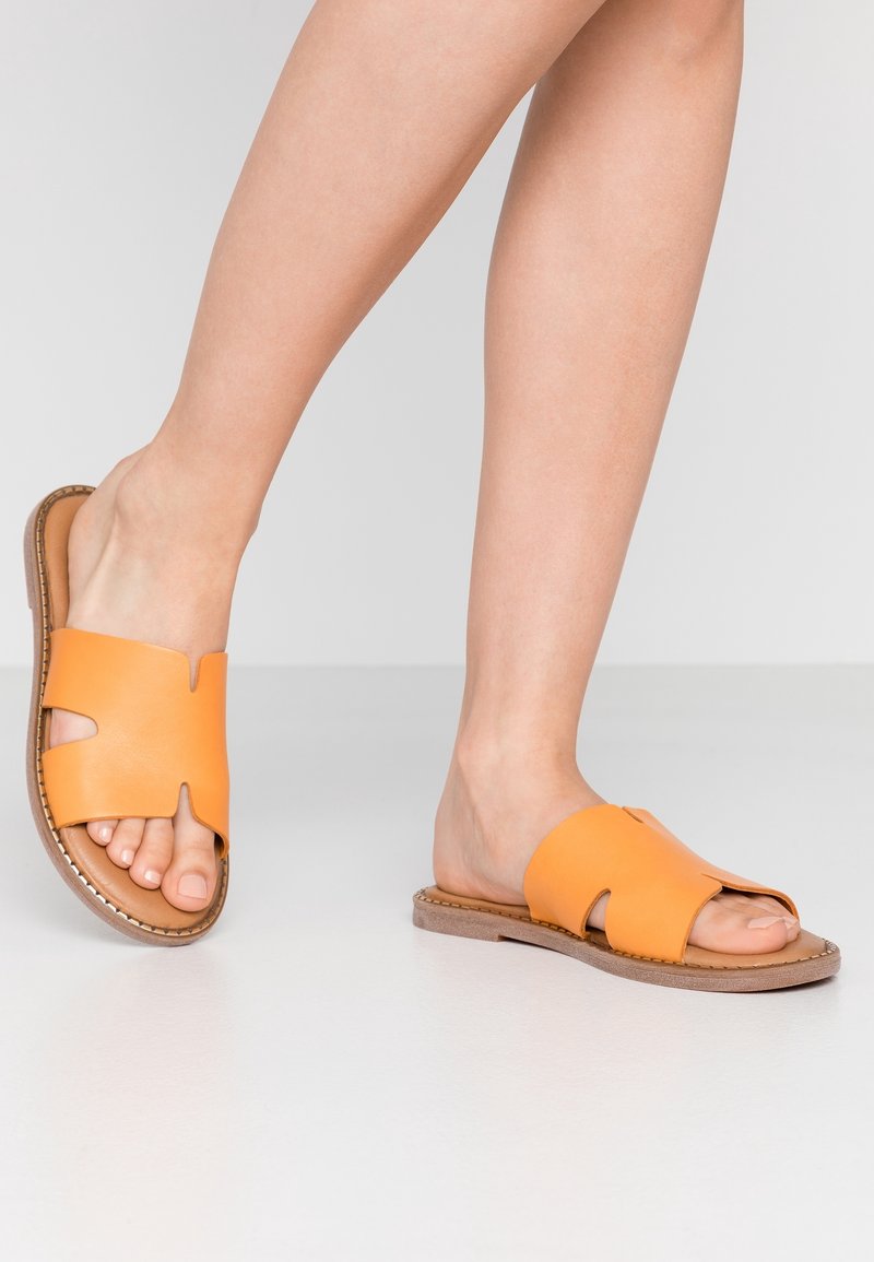 Sandales mule à bout ouvert en cuir orange vif. Présentent un design découpé avec une semelle plate marron et des coutures contrastantes.